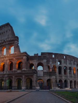 Imagem do Coliseu de Roma, uma estrutura circular com diversos arcos em suas entradas e janelas dispostas por toda a estrutura, feita de tijolos acinzentados teto aberto.